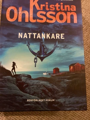 Nattankare av Kristina Ohlsson - Spännande skönlitterär bok med titeln 'Nattankare' av Kristina Ohlsson. Omslaget har en dramatisk illustration med ett stort ankare, en siluett av en person, hav, klippor och röda sjöbodar i bakgrunden. Perfekt för dig som gillar mysterier och äventyr.