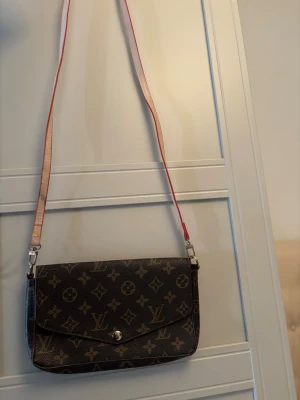 Kuvertväska - Stilren kuvertväska med klassiskt monogrammönster i brunt och beige. Väskan har en avtagbar axelrem i ljusrosa med röda detaljer och silverfärgade metalldelar. Perfekt för att bära det viktigaste med en lyxig touch.