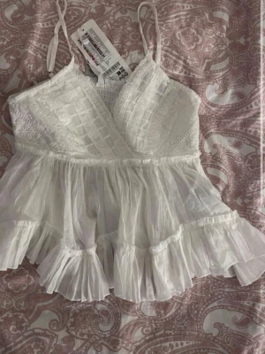 Vit volang topp med spetsdetaljer - Supersöt vit linnetopp med tunna axelband, omlott framtill och broderade spetsdetaljer. Toppen har volangkant och luftig passform, perfekt för varma dagar. Ger en romantisk och bohemisk vibe.