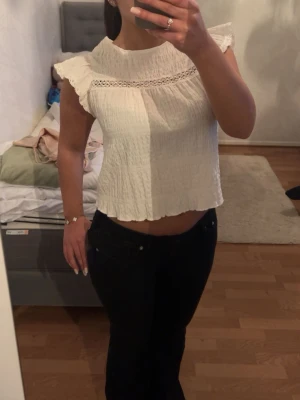 Vit croppad blus med spetsdetalj - Säljer en söt vit croppad blus med volangärmar och spetsdetalj vid halsen. Toppen har en lätt skrynklig struktur och är perfekt för dig som gillar en romantisk och trendig stil.