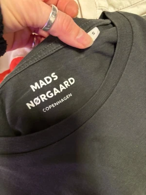 Svart t-shirt från Mads Nørgaard stl S - Snygg svart t-shirt från Mads Nørgaard Copenhagen i bomull. Klassisk rund hals och korta ärmar. Diskret detalj med svartvit randig etikett vid nederkanten. Perfekt basplagg med stilren look.