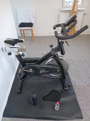 Svart spinningcykel med rem med tillbehör - Säljer en svart spinningcykel med remmar och inte kedja för tysthet när man trampar med digital display, justerbart styre och sadel. Medföljer vattenflaska, sadelöverdrag, svett skyddsmatta och lite olja på flaska för insmorning av remmarna. Maxvikt som kan sitta på cykeln är 120 kilo. Själva hjulet är på 20 kilo. Perfekt för hemmaträning och kondition. Men också toppen vara för professionella tränings anläggningar. Allt är i fungerande skick utan några som helst rörelsefel!