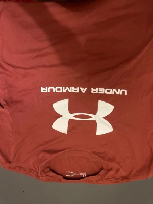 Röd Under Armour t-shirt - Säljer en röd t-shirt från Under Armour med stor vit logga och text på bröstet. T-shirten har en klassisk passform och är tillverkad i ett mjukt material som passar perfekt till träning eller chill. Rund halsringning och korta ärmar.