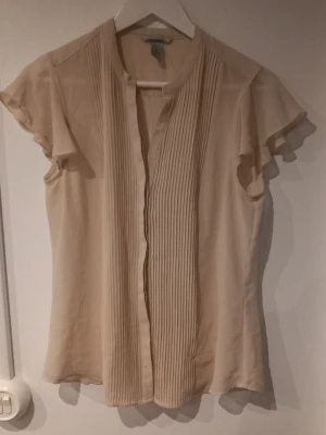Puderrosa blus med plisserad front H&M - Stilren puderrosa från H&M med plisserade detaljer framtill och korta, fladdriga ärmar. Blusen har en rak passform och rundad halsringning. Perfekt att matcha med både jeans och kjol för en trendig look.