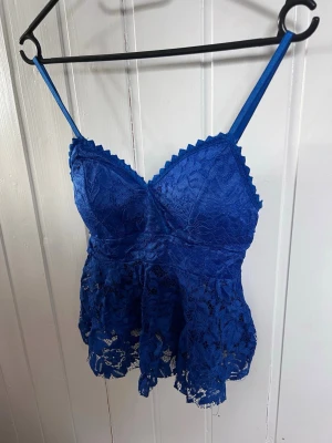 Blå spetsblus med smala axelband - Snygg blå blus i spets med smala axelband och v-ringning. Blusen har figurnära passform upptill och ett lätt utsvängd nederdel med transparent spetsdetalj. Perfekt för dig som vill ha en trendig och feminin look.