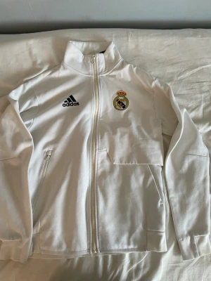 Real Madrid Adidas - Zip up - (Pris kan diskuteras) Snygg vit träningsjacka från Adidas med Real Madrid-logga på bröstet. Jackan har hel dragkedja, hög krage och två fickor med dragkedja. Tillverkad i ett mjukt och sportigt material, perfekt för fotbollsträning eller chill.