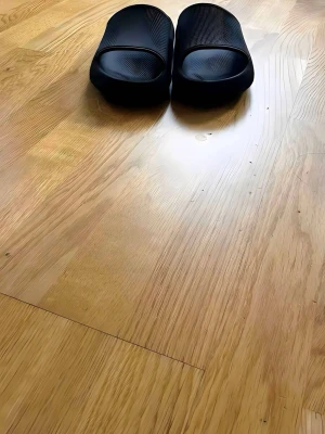 Svarta yeezy tofflor från addidas - Säljer ett par svarta chunky tofflor med öppen tå och ribbad yta. Tillverkade i mjukt gummi för extra komfort och en modern look. Perfekta för att glida runt hemma eller på gymmet. Snygg minimalistisk design utan synligt märke.
