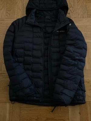 Svart dunjacka Peak Performance - Säljer en svart dunjacka från Peak Performance med huva och diskret logga på bröstet och ärmen. Jackan har quiltad design, dragkedja framtill och är perfekt för kalla vinterdagar. Materialet är lätt och värmande dun som håller dig varm hela säsongen. Den har två små hål längst ner som syns knappt