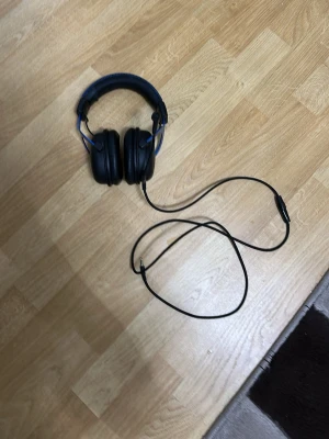 Svarta over-ear hörlurar med mikrofon - Säljer ett par svarta over-ear hörlurar med blå detaljer och vadderad bygel. Hörlurarna har en fast mikrofon och lång sladd med 3,5 mm kontakt. Perfekta för gaming eller musiklyssning. Materialet är mestadels plast och konstläder.