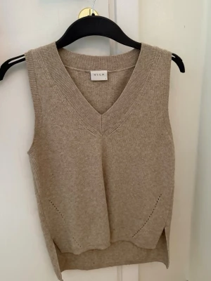Beige stickad väst från Vila - Beige stickad väst från Vila med v-ringning och diskreta fickdetaljer framtill. Västen har en något längre bakdel och ribbade kanter för en snygg finish. Perfekt att styla över en skjorta eller t-shirt.