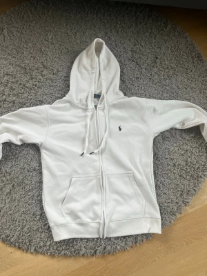 Vit hoodie från Polo Ralph Lauren - Säljer en clean vit hoodie från Polo Ralph Lauren med dragkedja, huva med snören och två stora fickor fram. Klassisk logga broderad på bröstet. Perfekt för dig som gillar stilrena och bekväma plagg. Materialet känns mjukt och skönt mot huden.