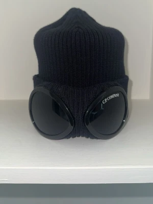 Svart Goggle mössa från C.P. Company - Cool svart ribbstickad mössa från C.P. Company med ikoniska svarta goggle-linser på uppviket. Mössan är tillverkad i mjukt stickat material och har en modern, urban look som sticker ut. Perfekt för dig som vill ha något unikt och trendigt.