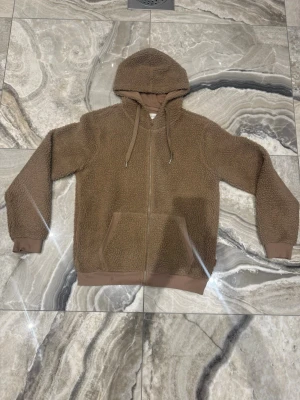 Brun teddyjacka med huva - Mysig brun teddyjacka med dragkedja framtill och stor huva. Jackan har två fickor fram, ribbade muddar vid ärmslut och nederkant samt dragsko i huvan. Perfekt för kalla dagar när du vill vara både varm och stilren.