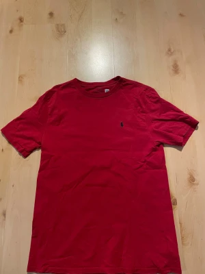 Röd t-shirt från Polo Ralph Lauren - Klassisk röd t-shirt från Polo Ralph Lauren med rund halsringning och den ikoniska broderade loggan på bröstet. Tillverkad i mjuk bomull och har en normal passform. Perfekt basplagg för en clean och stilren look.