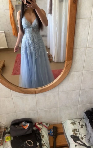 Ljusblå långklänning med spetsdetaljer - Elegant ljusblå långklänning med v-ringning och tunna axelband. Klänningen har vackra spetsdetaljer upptill och ett lager av tunn tyll som ger en drömmig look. Perfekt för bal eller student. Öppen rygg och figurnära passform upptill.