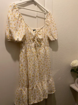 Blommig kortklänning Gina Tricot - Supersöt kortklänning från Gina Tricot i vitt med små gula och orange blommor. Klänningen har puffärmar, snörning framtill och volang nedtill. Perfekt för sommarens alla tillfällen och har en luftig, lätt känsla.