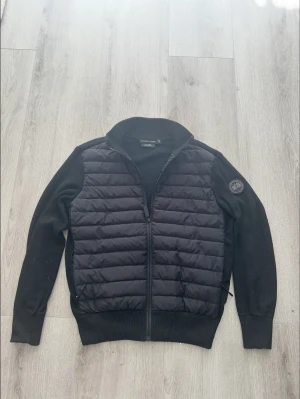 Svart cardigan från canada goose - Snygg svart cardiganjacka från Canada goose med vadderad framsida och stickade ärmar samt rygg. Jackan har dragkedja framtill, ribbade muddar och en patch på ena ärmen. Perfekt för lager-på-lager och streetwear-stil. Står M passar Som S