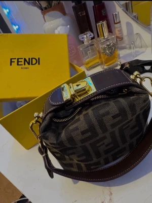 Fendi handväska  - Säljer en snygg handväska med klassiskt FF-mönster i brunt och svart. Väskan har ett brett brunt läderhandtag, guldfärgade metalldetaljer och en låsdetalj framtill. Medföljer originalkartong och extra axelrem.