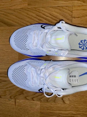 Vita Nike sneakers med blå detalj - Snygga vita Nike sneakers med svart swoosh och blå detaljer på sulan. Ovandelen är i ventilerande mesh och skorna har klassisk snörning. Perfekta för dig som gillar sportig stil och vill ha bekväma skor till vardags. Lätta och stilrena med modern design.