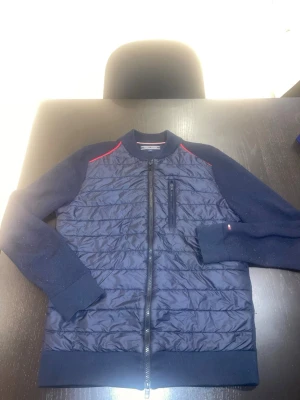 Marinblå bomberjacka Tommy Hilfiger - Snygg marinblå bomberjacka från Tommy Hilfiger med quiltad front, stickade ärmar och muddar. Jackan har en bröstficka med dragkedja, diskret logga på ärmen och röda detaljer vid axlarna. Perfekt för dig som vill ha en sportig och stilren look.