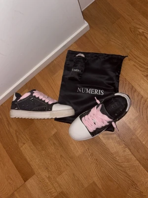 Numeris skor med rosa snören - Snygga låga sneakers från Numeris i svart och vitt med rosa skosnören. Skorna har en vit tå och häl,  ovandel i jeans med stilrena slitningar och en klassisk gummisula. Perfekta för dig som vill sticka ut med färgglada snören och stilren design.