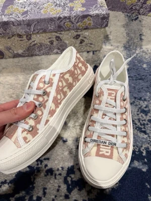Rosa Dior sneakers med monogram - Snygga sneakers från Christian Dior i ljusrosa tyg med ikoniskt Dior-monogram. Skorna har vita gummisulor, vita snören och en tydlig logga på plösen. Sulan har en svart stjärndetalj undertill. Perfekta för dig som vill ha en exklusiv och trendig look.