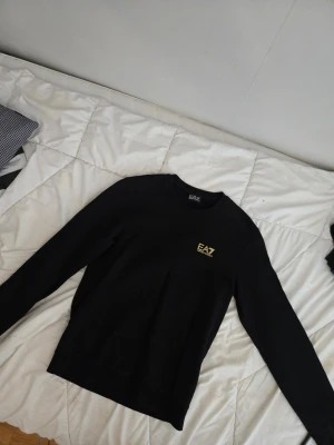 Svart sweatshirt från EA7 Emporio Armani - Aldrig använd, Svart sweatshirt med rund hals från EA7 Emporio Armani. Tröjan har en gul EA7-logga på bröstet och guld detalj i nacken. Långärmad modell i mjukt material, perfekt för en clean och stilren look.