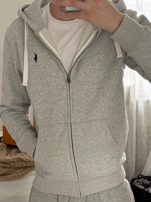 Grå zip hoodie från Ralph Lauren - Snygg ljusgrå hoodie med dragkedja från Ralph Lauren. Klassisk design med huva och vita dragsnören, ribbade muddar och fickor framtill. Ikoniska blå loggan broderad på bröstet. Perfekt för en avslappnad och stilren look.