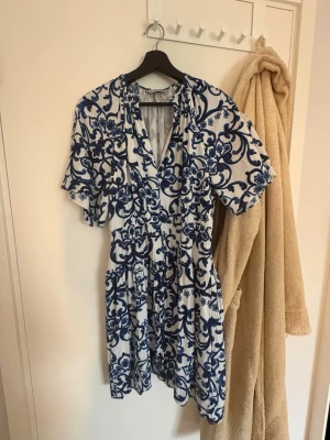 Vit och blå kortklänning med blommönster - Kortklänning i vitt med blått blommönster och v-ringning. Klänningen har korta ärmar och markerad midja för en feminin siluett. Perfekt för varma dagar och har en luftig känsla. Tillverkad i ett mjukt bomullsliknande material.