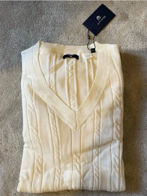 Vit tröja från GANT - Vit tröja från GANT med djup V-ringning och ribbade muddar. Tröjan har ett klassiskt mönster med flätade detaljer och är tillverkad i mjukt material som känns skönt mot huden. Perfekt för dig som gillar stilrena och tidlösa plagg.