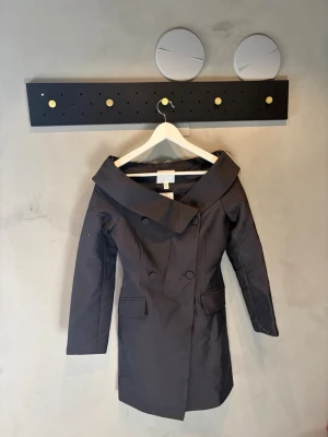 Mörkgrå/svart trenchcoat från H&M - Stilren mörkgrå trenchcoat från H&M med bred krage och dubbelknäppning framtill. Jackan har två fickor med lock och en figurnära passform som ger en snygg siluett. Perfekt för dig som vill ha en klassisk men modern look.