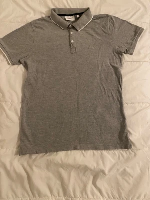 Grå pikétröja från Lindbergh - Klassisk grå pikétröja från Lidbergh med vit kant på kragen och tre knappar framtill. Tröjan har korta ärmar och en stilren look som passar till det mesta. Perfekt för dig som gillar enkel och tidlös stil.