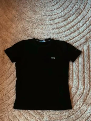 Svart t-shirt från Lacoste - Klassisk svart t-shirt från Lacoste med den ikoniska krokodilloggan broderad på bröstet. T-shirten har rund halsringning och korta ärmar. Perfekt basplagg som funkar till allt och är riktigt skön att bära.