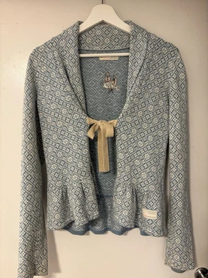 Blåmönstrad kofta med rosett - Supersöt blå och vit kofta från Odd Molly med diskret diamantmönster, volangkant nedtill och beige rosett framtill. Långärmad modell med broderad detalj på ryggen. Perfekt för dig som gillar unika detaljer och skön passform.
