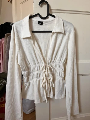 Vit ribbad topp med knyt från Gina Tricot - Vit långärmad topp från Gina Tricot i ribbat material. Djup v-ringning, krage och snygga knytband framtill som ger en figurnära passform. Perfekt för att skapa en trendig och avslappnad look.