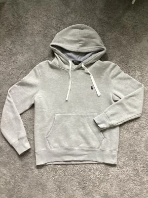 Grå hoodie från Polo Ralph Lauren - Snygg ljusgrå hoodie från Polo Ralph Lauren med klassisk huva och vita snören. Broderad lila logga på bröstet och stor magficka framtill. Perfekt för chill dagar och enkel att matcha med jeans eller joggers.