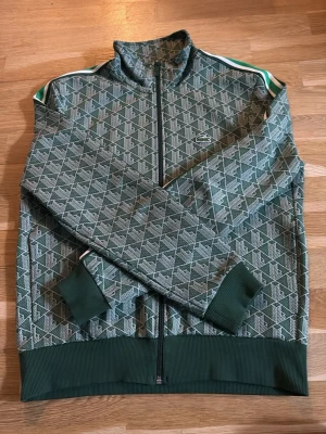 Grön mönstrad ziptröja från Lacoste - Snygg grön ziptröja från Lacoste med vitt geometriskt mönster och klassisk krokodil-logga på bröstet. Tröjan har ribbade muddar, hög krage och vita/gröna ränder på axlarna. Perfekt för en sportig och trendig look.