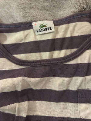 Randig långärmad topp från Lacoste - Säljer en långärmad topp från Lacoste med breda lilla och vita ränder. Toppen har rund halsringning och en liten bröstficka med den klassiska Lacoste-loggan. Tillverkad i mjuk bomull, perfekt för en avslappnad stil.