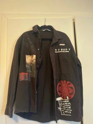 Svart overshirt med tryck och patchar - Unik svart overshirt med vita tryck, röda och svarta patchar samt texten 'NOBODY' på ryggen och rygg. Framsidan har konstnärliga detaljer och grafiska motiv.  Knappstängning och en bröstficka. Perfekt för dig som gillar streetwear och statement-plagg.
