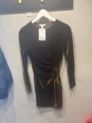 Olivgrön sammetsklänning från H&M - Snygg olivgrön sammetsklänning från H&M med lång ärm och omlottdetalj i midjan. Klänningen har en kroppsnära passform och är i kort modell, perfekt för dig som gillar en elegant och trendig look.