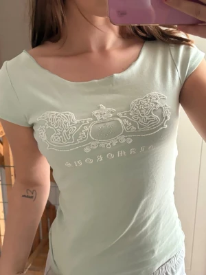 Ljusgrön topp med tryck  - Snygg ljusgrön topp  med korta ärmar och ett ikoniskt vitt tryck framtill. T-shirten har en figurnära passform och är gjord i mjuk bomull som känns skön mot huden. Perfekt för dig som gillar en enkel men trendig stil.