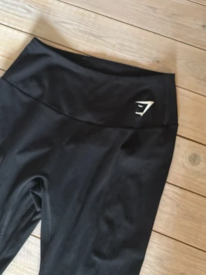 Svarta leggings från Gymshark - Snygga svarta leggings från Gymshark med hög midja och diskret logga framtill. Tillverkade i stretchigt material som sitter skönt och formar sig efter kroppen. Perfekta för träning eller chill dagar.