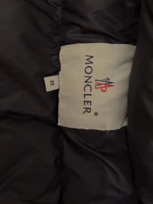 Svart dunjacka från Moncler - Snygg svart dunjacka från Moncler med klassisk quiltad design. Jackan har en ikonisk Moncler-logga på insidan och ett färgglatt serietidningsfoder. Perfekt för kalla vinterdagar och riktigt varm tack vare dunfyllningen.