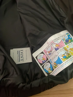Svart dunjacka från Moncler - Snygg svart dunjacka från Moncler med klassisk logotyp på ärmen och coolt serietryck på insidan. Jackan har en stilren design och är fylld med dun för maximal värme under kalla dagar. Perfekt för dig som vill ha både stil och funktion i vinter.