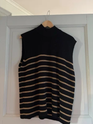 Randig stickad väst med polokrage - Säljer en svart stickad väst med beigea horisontella ränder och hög polokrage. Västen är ärmlös och har en loose passform, perfekt att styla över en skjorta eller t-shirt för en trendig lager-på-lager-look.