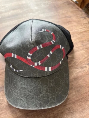 Gucci keps  - Säljer en svart och grå keps från Gucci med klassiskt GG-mönster och en röd, vit och svart orm på framsidan. Kepsen har mesh på baksidan för extra ventilation och justerbar passform. Perfekt för dig som vill sticka ut med en lyxig accessoar. Knoppen på toppen är borta. Spännet är slitet men ingen man tänker på när den används. Köpt på vinted! 