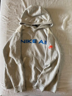 Grå Nike Air hoodie med blå logga - Snygg grå hoodie från Nike Air med stor blå logga på bröstet och röd Nike Air-detalj på ärmen. Hoodien har huva, vita axelpartier och en klassisk magficka. Tillverkad i mjukt sweatshirtmaterial som är perfekt för chill dagar.