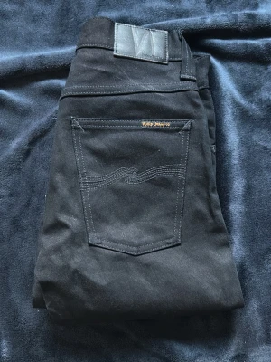 Svarta jeans från Nudie Jeans - Säljer ett par svarta jeans från Nudie Jeans med klassisk femficksmodell och diskret broderad bakficka. Jeansen har raka ben och är tillverkade i ett slitstarkt bomullstyg. Perfekta för dig som gillar stilrena och tidlösa plagg.