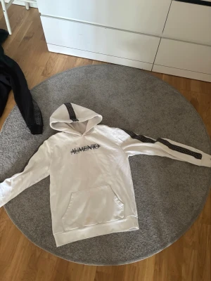 Vit hoodie från Aumento med tryck - Säljer en vit hoodie från Aumento med svart tryck på bröstet och dekorativa band längs ärmarna. Hoodien har en klassisk känguruficka framtill och huva. Perfekt för dig som gillar streetwear och vill ha en stilren men ändå unik look.