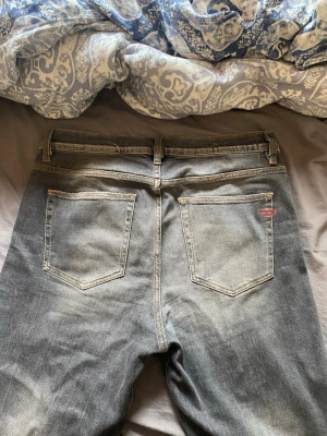 Grå slitna jeans från Diesel - Säljer ett par grå jeans från Diesel med slitningar framtill och klassisk femficksmodell. Jeansen har en tvättad look och raka ben. Märkets logga syns på fickan. Perfekta för en avslappnad och trendig stil.